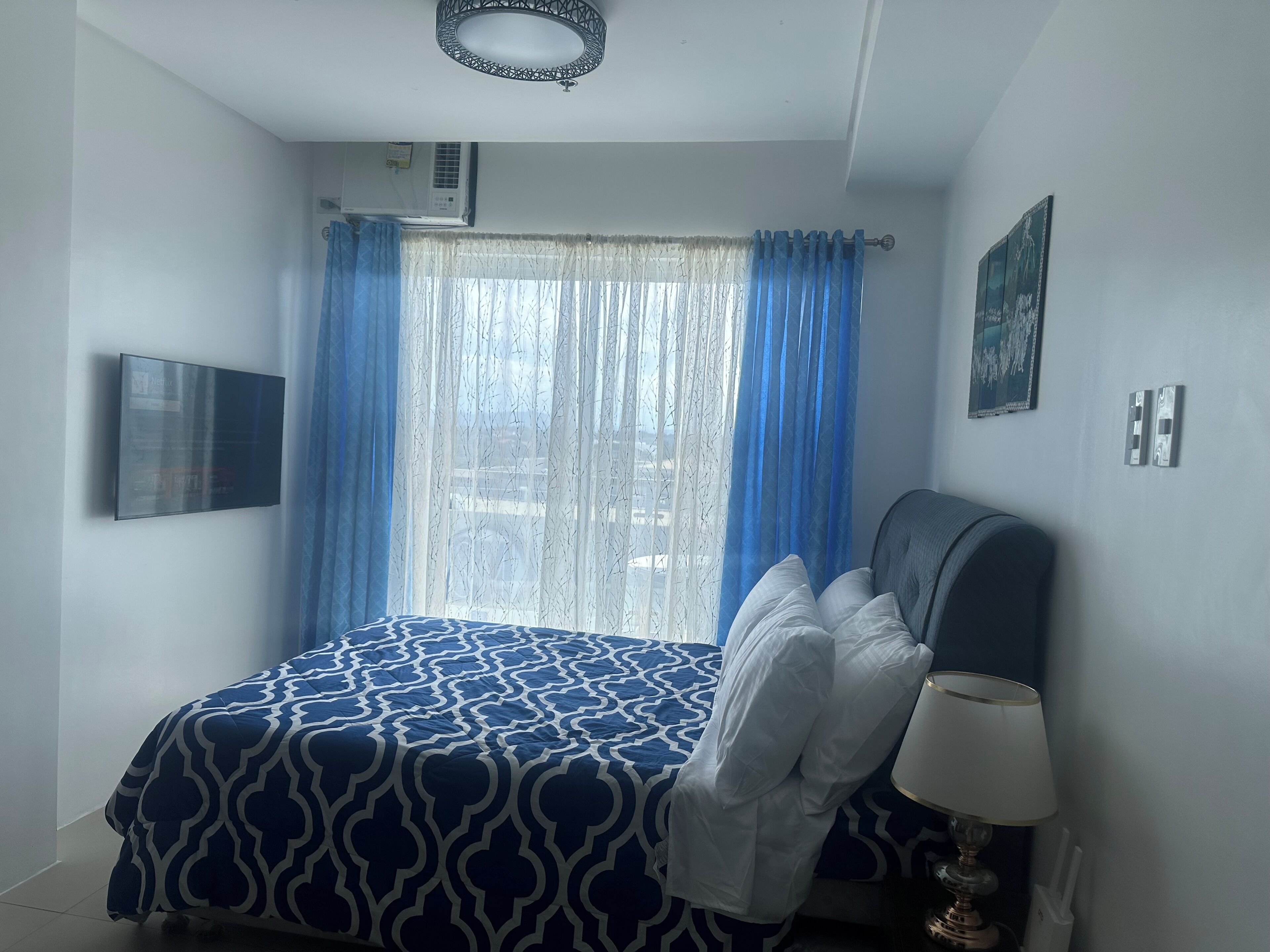 A bedroom with a bed, a TV mounted on the wall, a window with blue curtains, and a wall-mounted air conditioner.