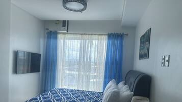A bedroom with a bed, a TV mounted on the wall, a window with blue curtains, and a wall-mounted air conditioner.