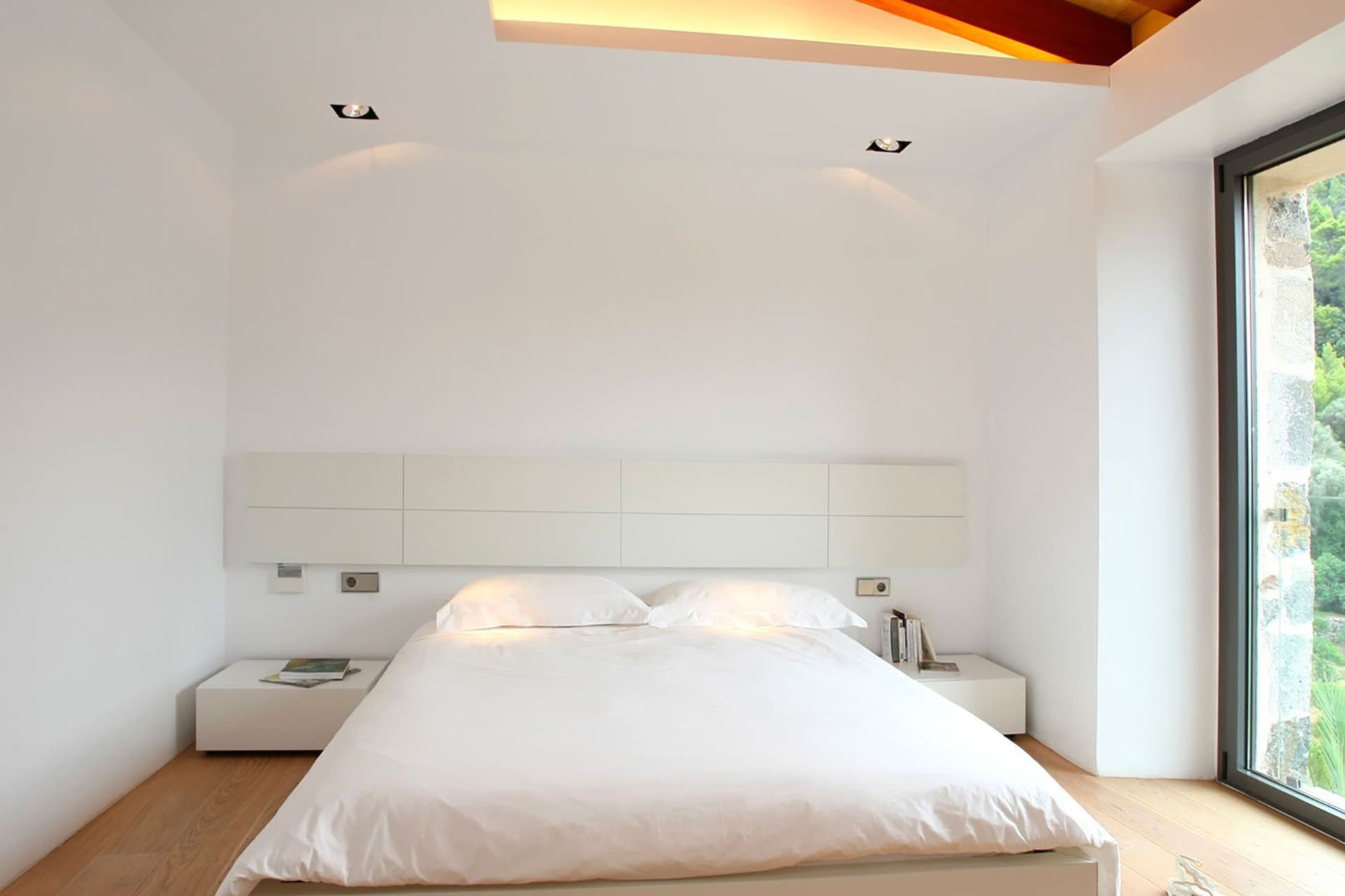 A modern bedroom with a large bed, bedside tables, and a view of greenery through a window.