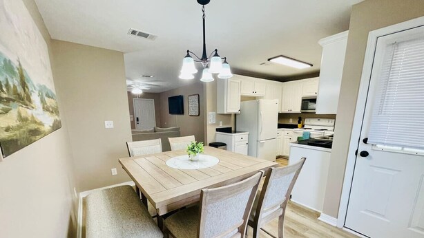 Un coin repas avec une table et des chaises en bois, un lustre et une cuisine en arrière-plan.