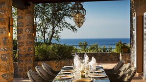 A dining table set for six with wicker chairs, overlooking a scenic view of the sea.