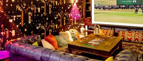 Cómoda zona de estar con un sofá de cuero, una mesa de centro con revistas y una pantalla grande mostrando un evento de golf.