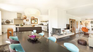 Una cocina moderna con una isla central, un comedor con mesa de madera oscura y sillas color turquesa, y una sala de estar con chimenea y sillón naranja.