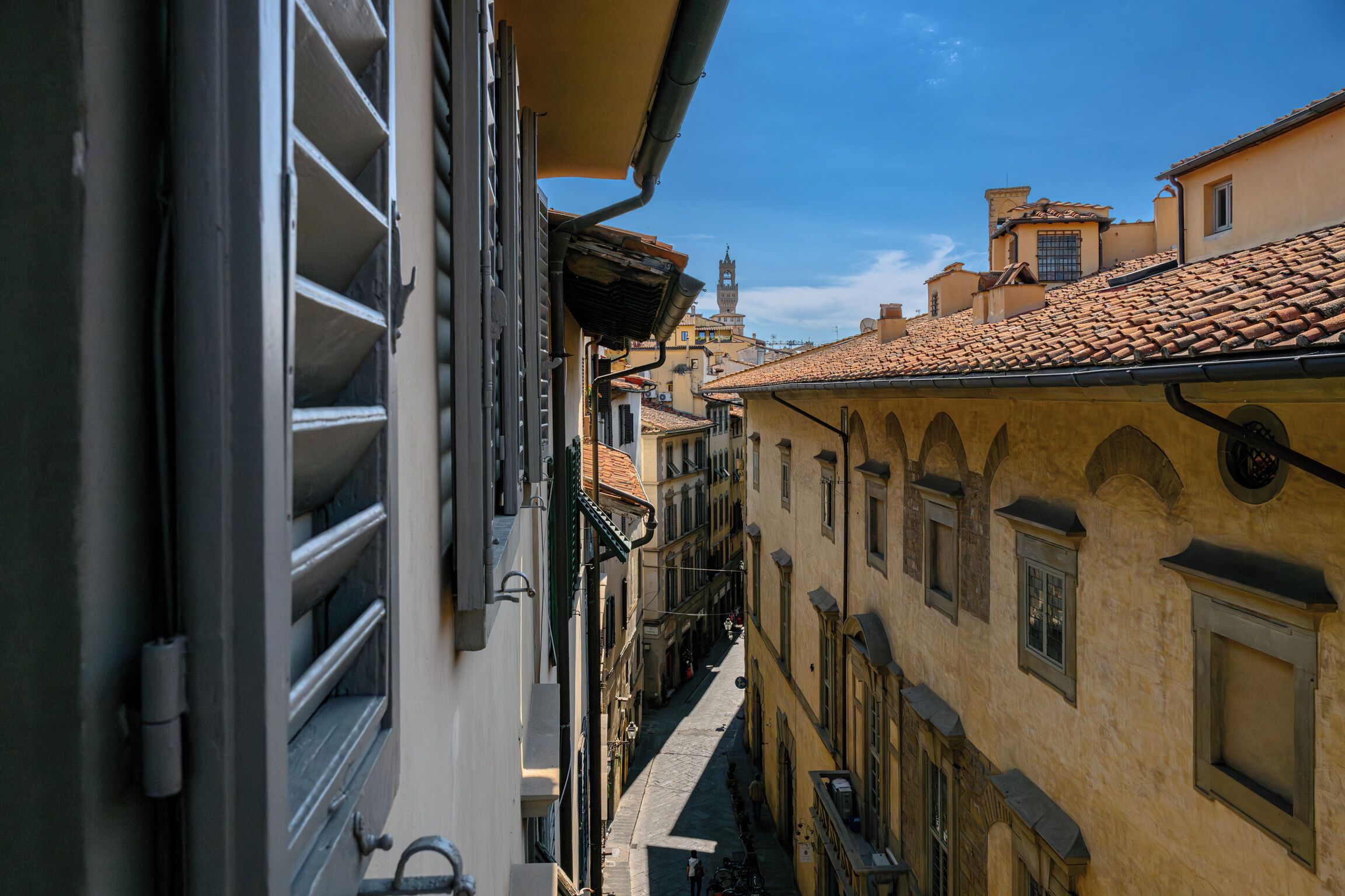 A narrow street with buildings on either side, featuring a mix of architectural styles and a clear blue sky.