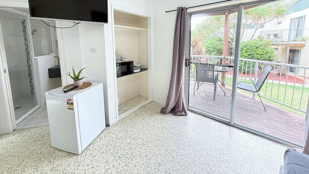 Un espace de vie compact comprenant un coin cuisine, un petit réfrigérateur, une plante en pot et une porte-fenêtre coulissante donnant sur une terrasse extérieure avec une table et des chaises.
