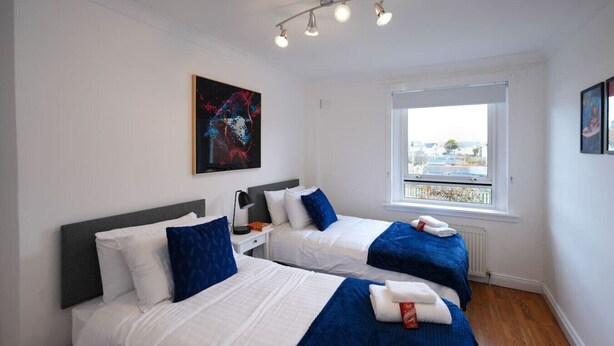 Ein Hotelzimmer mit zwei Betten, weißer Bettwäsche, blauen Zierkissen, einem Gemälde an der Wand und einem Fenster mit Blick auf die Stadt.
