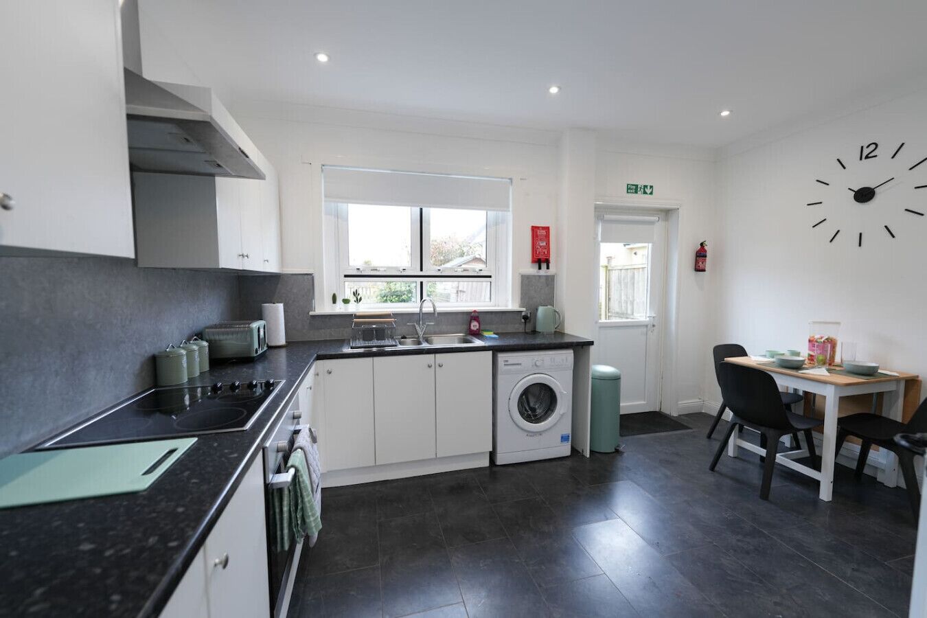 A modern kitchen with a stove, sink, and washing machine, a dining area with chairs, and a large wall clock.