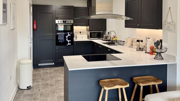 A modern kitchen with dark cabinetry, a central island with stools, and a stainless steel range hood. - Upper Townhouse, Exeter (Devon)