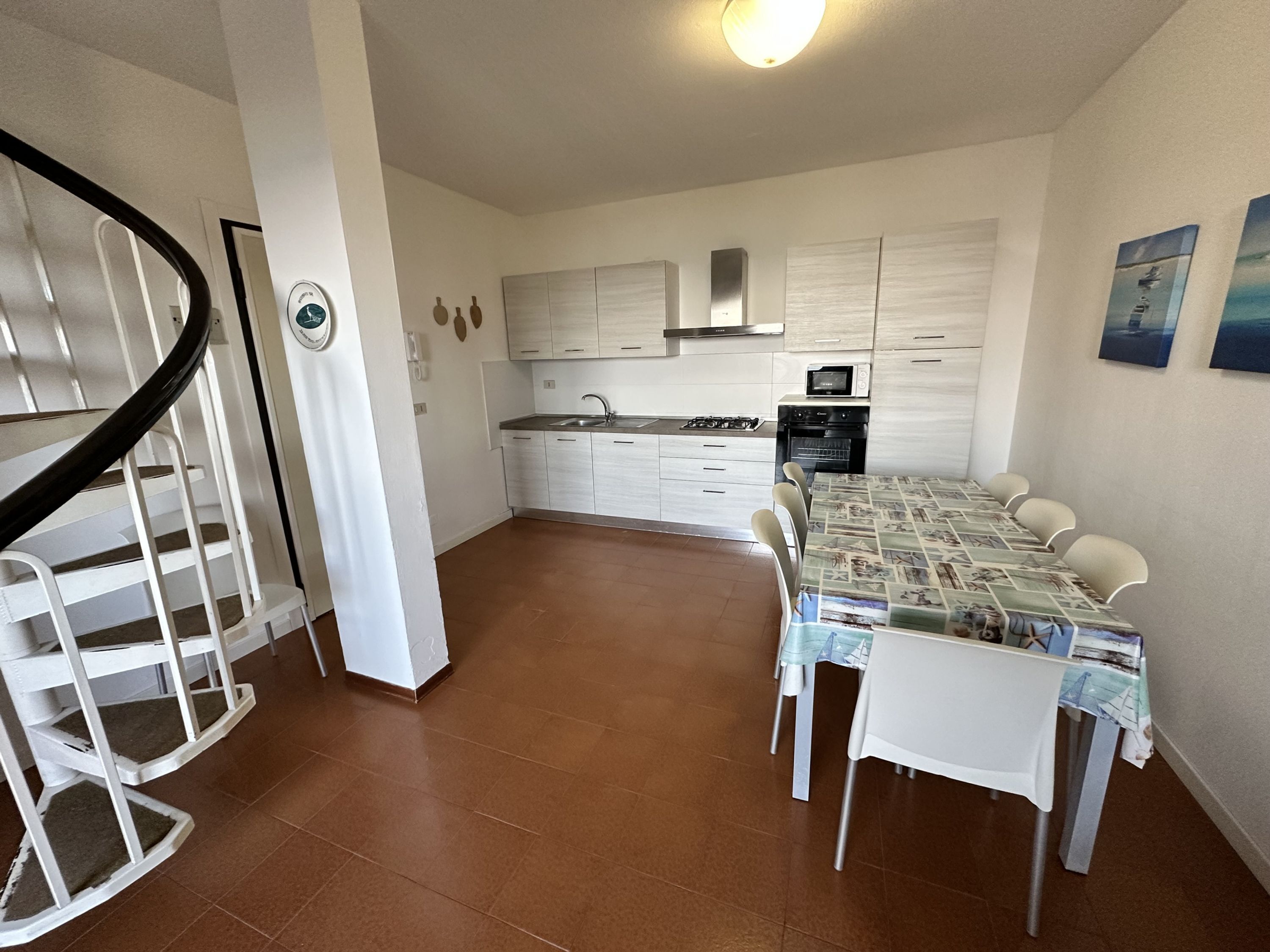 Una cocina moderna con armarios blancos, un microondas y una estufa a gas. Hay un comedor con una mesa y sillas, y una escalera con barandilla blanca.
