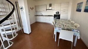 Eine moderne Küche mit weißen Schränken, einer Mikrowelle und einem Gasherd. Ein Essbereich mit Tisch und Stühlen sowie eine Treppe mit weißer Geländer sind ebenfalls vorhanden.