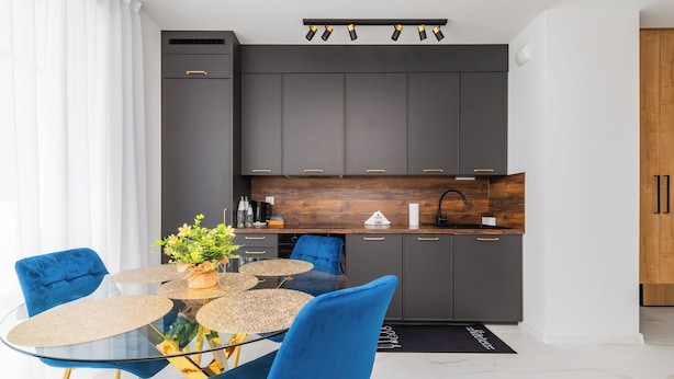 Une cuisine moderne avec des meubles de couleur sombre, un plan de travail en bois et une table à manger en verre, assortie de chaises bleues.