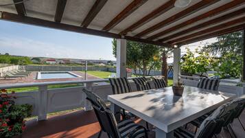 A covered outdoor patio with a table and chairs, a swimming pool, and a view of houses in the distance.
