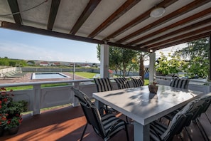 A covered outdoor patio with a table and chairs, a swimming pool, and a view of houses in the distance.