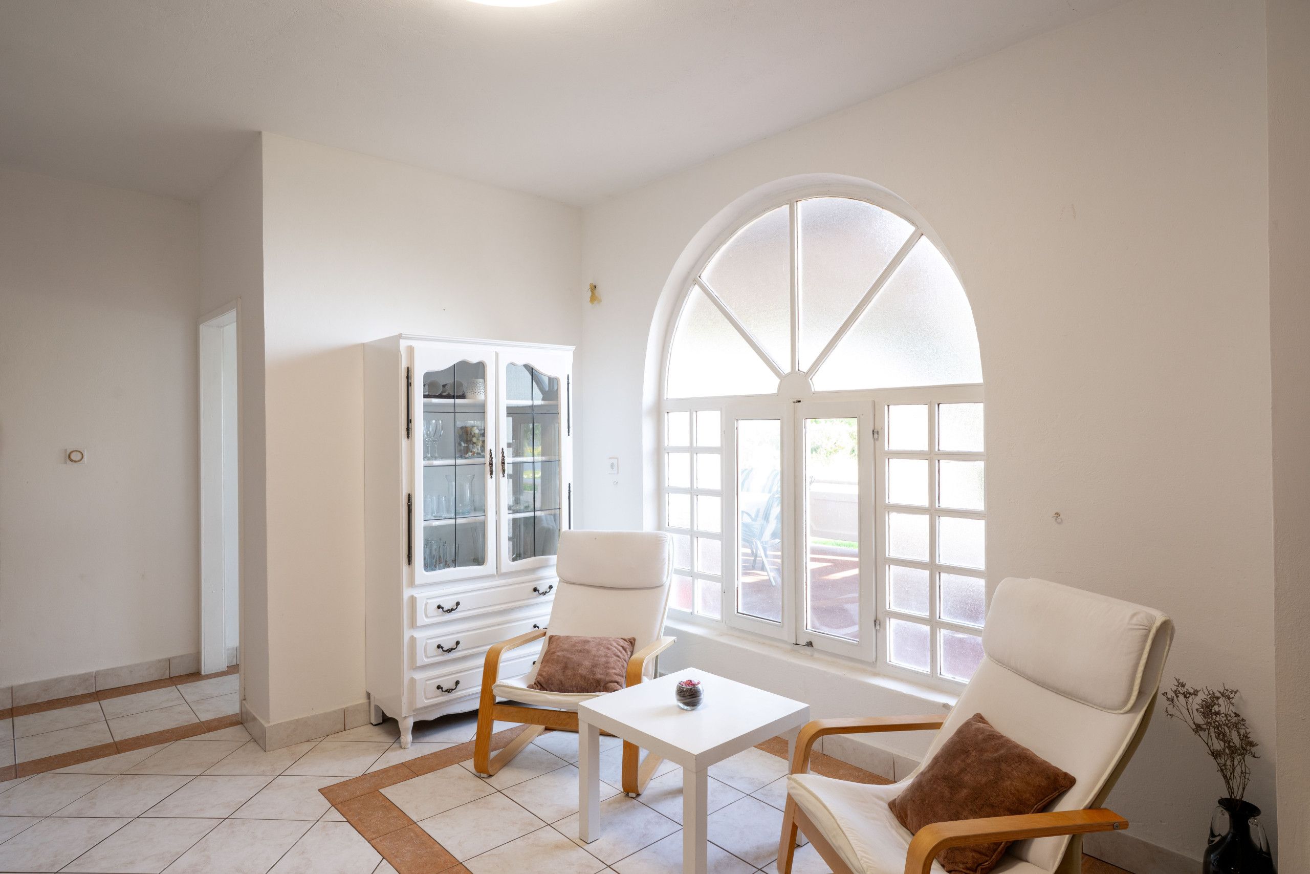 A room with a white cabinet, two chairs, a small table, and a large arched window.