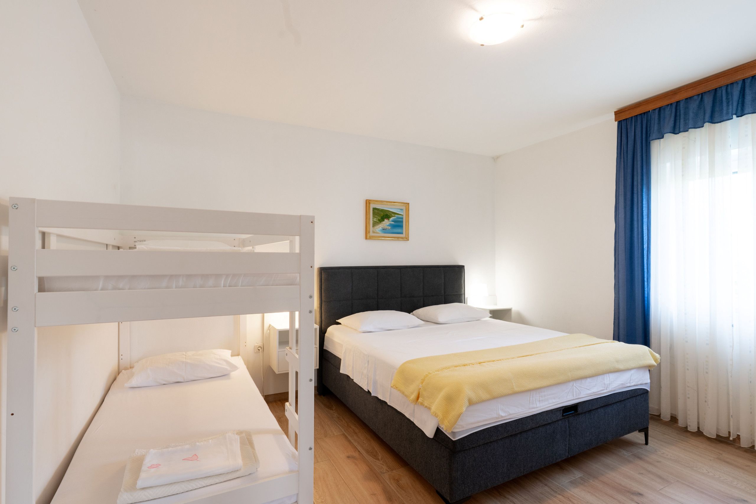 Une chambre d’hôtel avec un lit superposé, un lit simple, une table de chevet, une lampe et un tableau au mur.