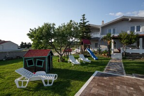 A residential area with a grassy lawn, a children's playhouse, a slide, and a pathway leading to a house.