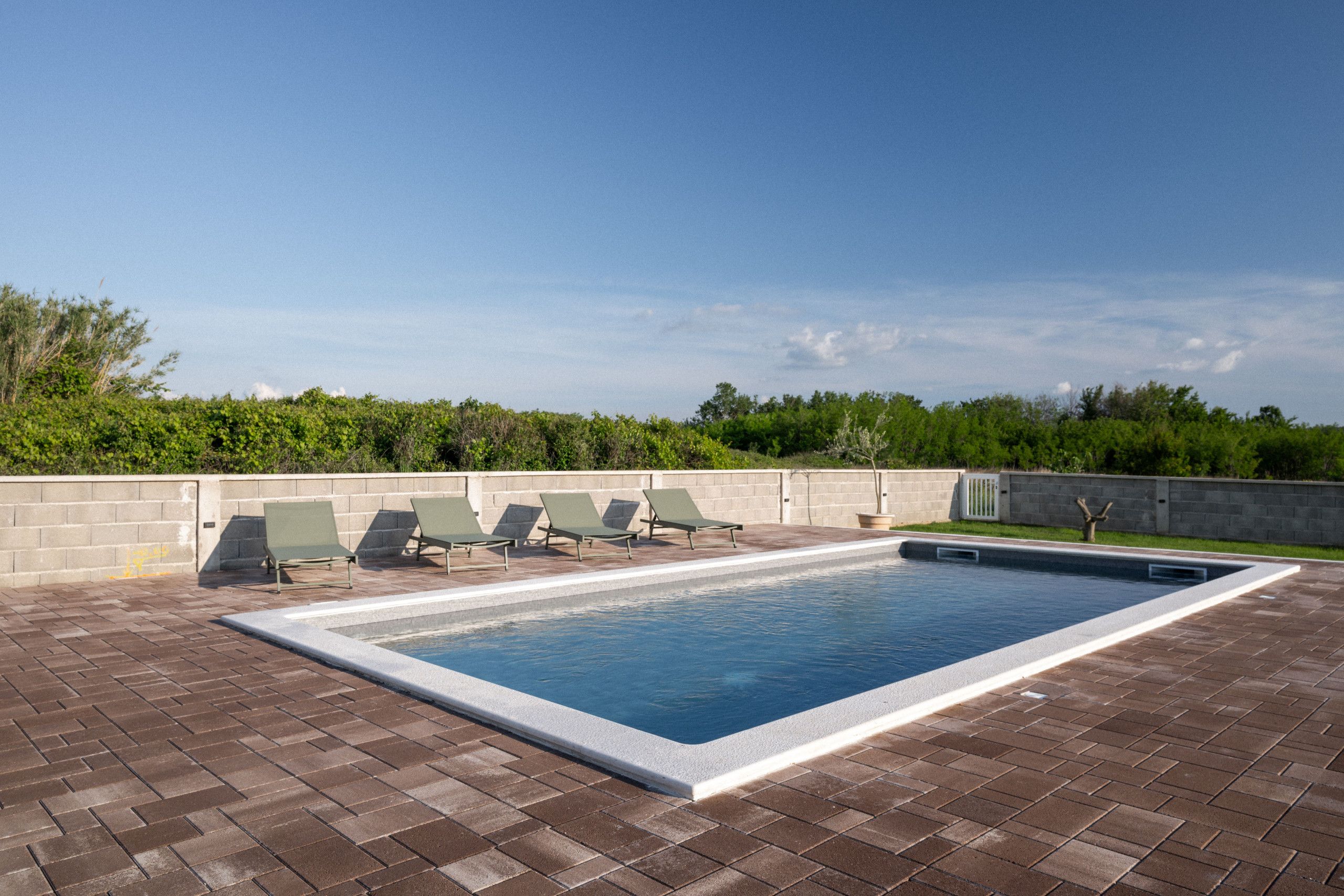 A rectangular swimming pool with a white border, surrounded by a brick patio. There are four green lounge chairs aligned along the pool's edge.