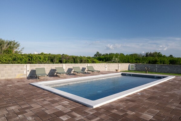 A rectangular swimming pool with a white border, surrounded by a brick patio. There are four green lounge chairs aligned along the pool's edge.