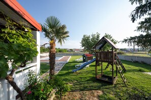 A playground with a slide and climbing structure, surrounded by a grassy area and a white building with a red roof.