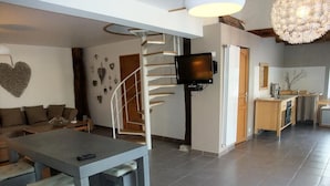 A modern living room with a grey sofa, a dining table with benches, a spiral staircase, a flat-screen TV mounted on the wall, and a kitchen area with a wooden countertop and cabinets.