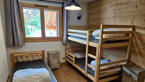 A dorm-style room with bunk beds, a single bed, a small bedside table, a lamp, and a window with curtains. - Bright apartment near slopes, pets allowed (Les Deux Alpes)