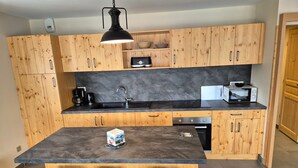 A modern kitchen with wooden cabinets, a black countertop, a built-in oven, a microwave, and a coffee maker. There are three wooden stools at the island. - Bright apartment near slopes, pets allowed (Les Deux Alpes)