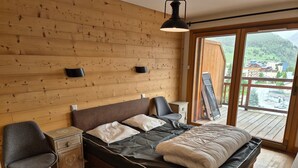 A bedroom with a wooden wall, a bed with a mattress and pillows, a chair, a nightstand, and a window with a view of a town. - Bright apartment near slopes, pets allowed (Les Deux Alpes)