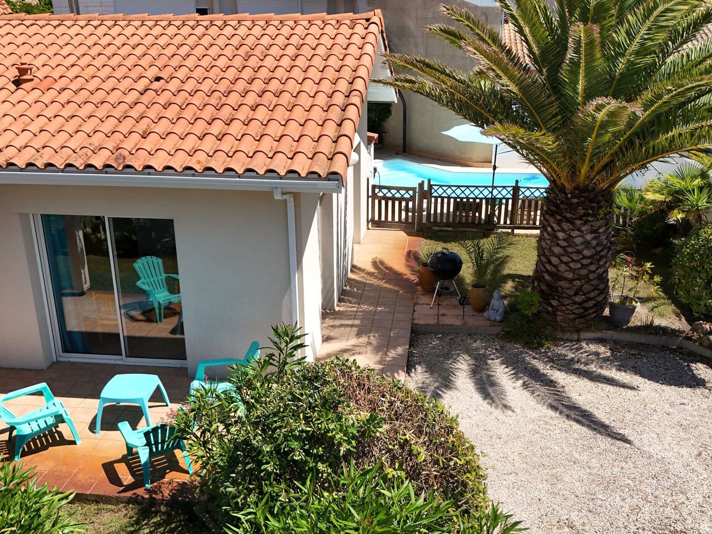 Een huis met een pannendak, een terras met stoelen, een barbecue en een palmboom.