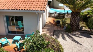 Een huis met een pannendak, een terras met stoelen, een barbecue en een palmboom.