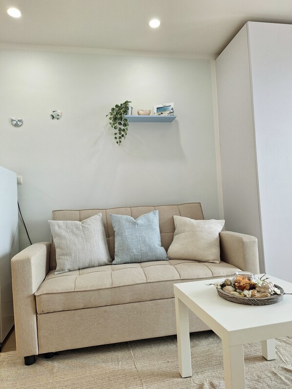 Un soggiorno dai toni neutri con un divano color beige, due cuscini, un tavolino con un oggetto decorativo e una mensola a parete con una pianta e due piccoli oggetti decorativi.
