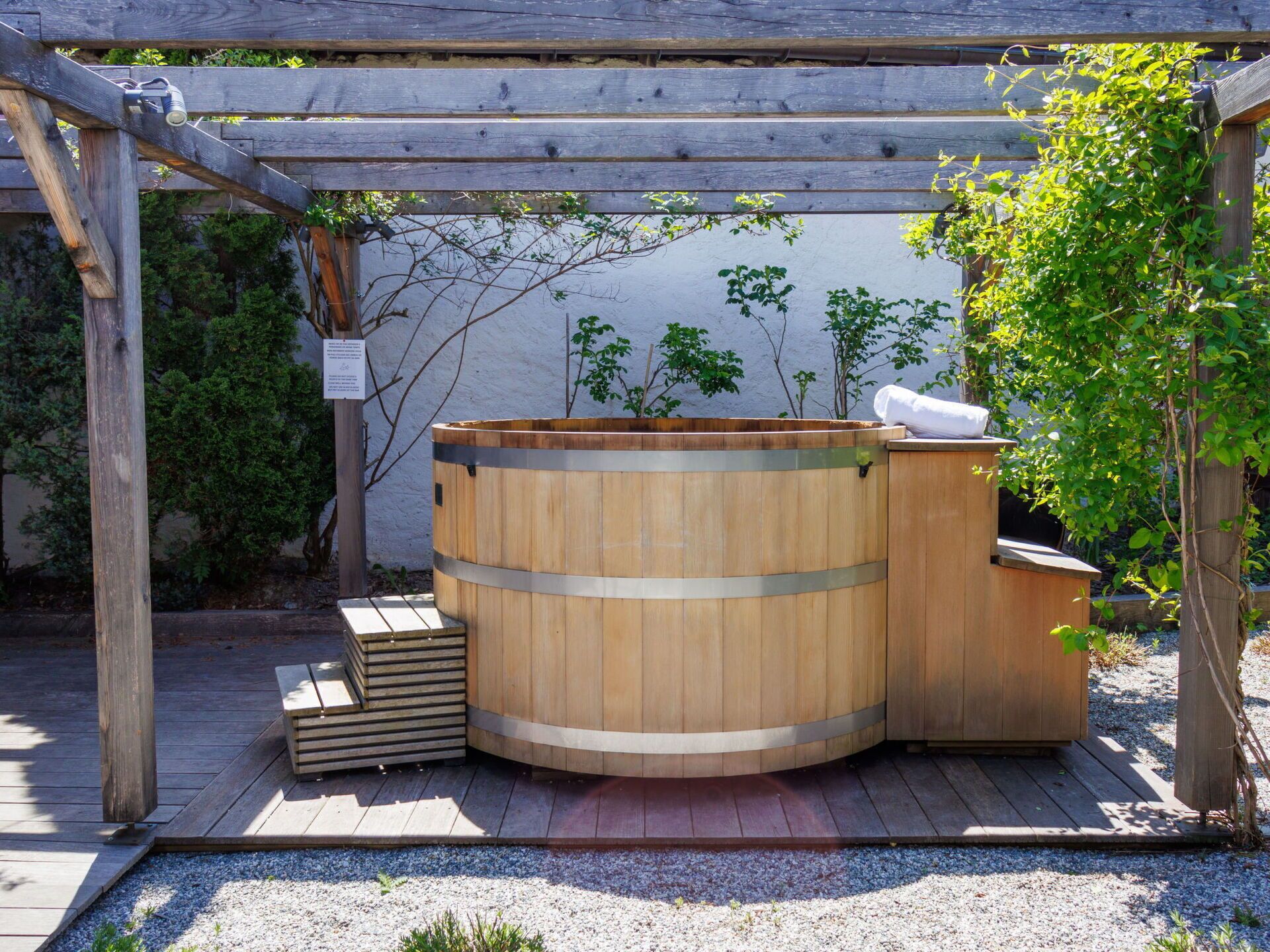 A wooden hot tub under a wooden pergola with a white wall and greenery in the background.