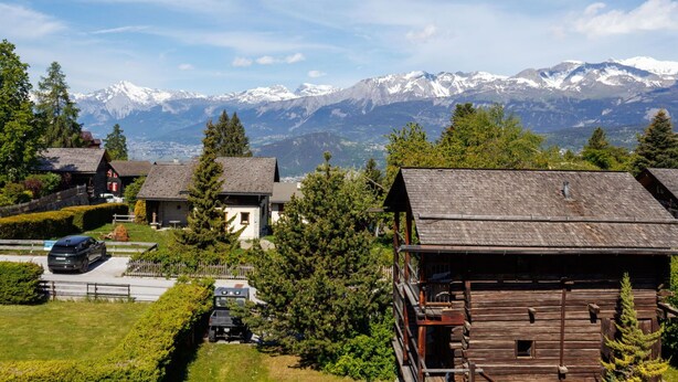 Ein Bergdorf mit traditionellen Holzhäusern, einer gepflasterten Auffahrt und Blick auf schneebedeckte Berge.