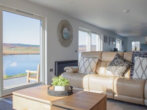 Amplia sala de estar con un gran ventanal con vista a un cuerpo de agua, un sofá de cuero color beige, una mesa de centro de madera y un espejo decorativo en la pared.