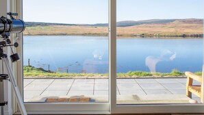 Une pièce avec un télescope monté sur un trépied, un banc et une vue sur un lac, avec des collines à l’horizon.