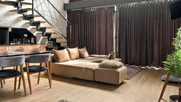 Un salon moderne avec un plancher en bois, un ensemble de canapés, un coin repas comprenant une table et des chaises, un escalier et une grande fenêtre avec des rideaux.