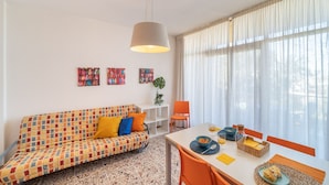 Una sala de estar moderna con un sofá colorido, una mesa de comedor con sillas naranjas y un amplio ventanal con cortinas translúcidas.