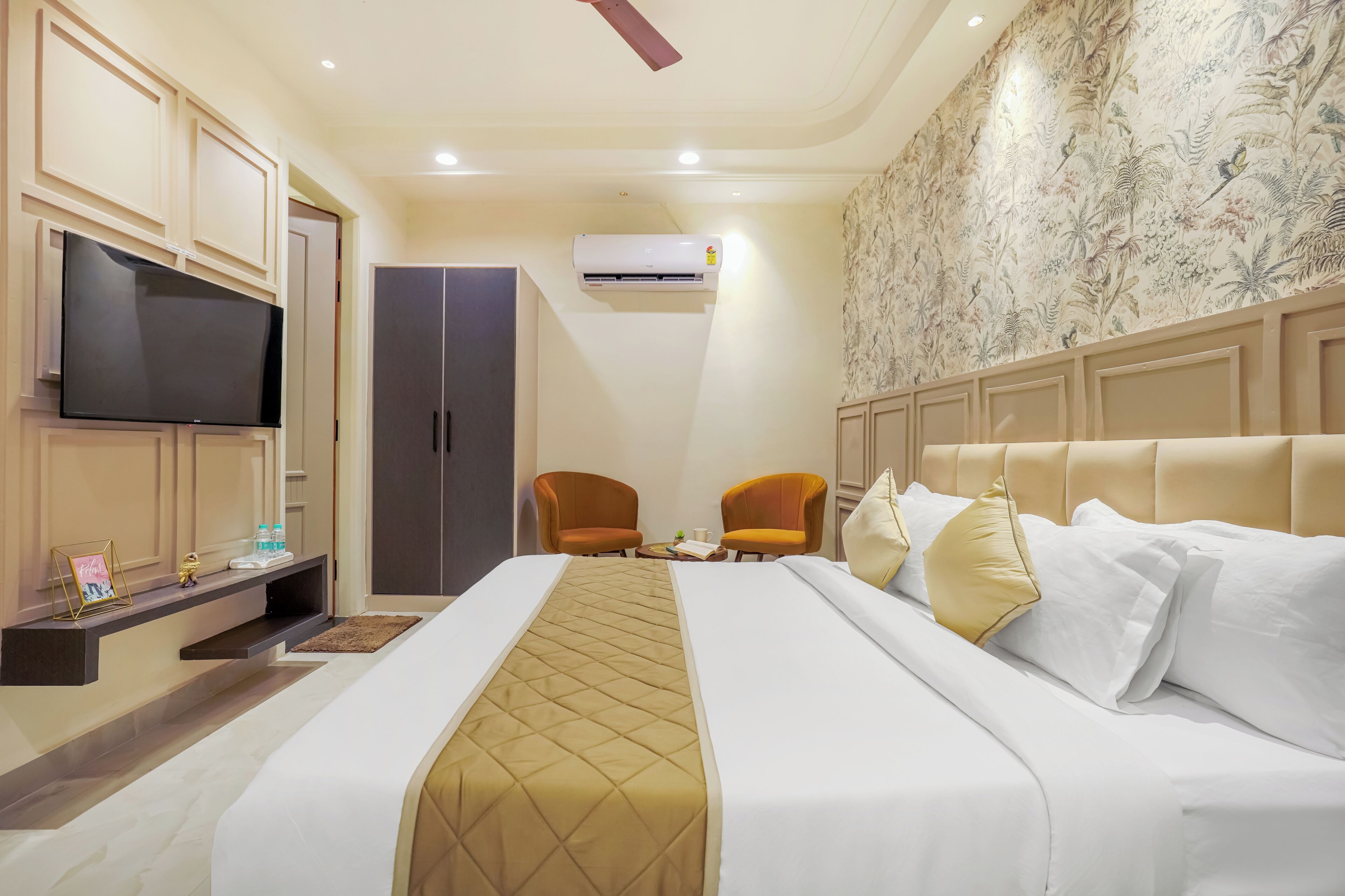 A hotel room with a bed, two chairs, a TV, a wardrobe, and an air conditioning unit.