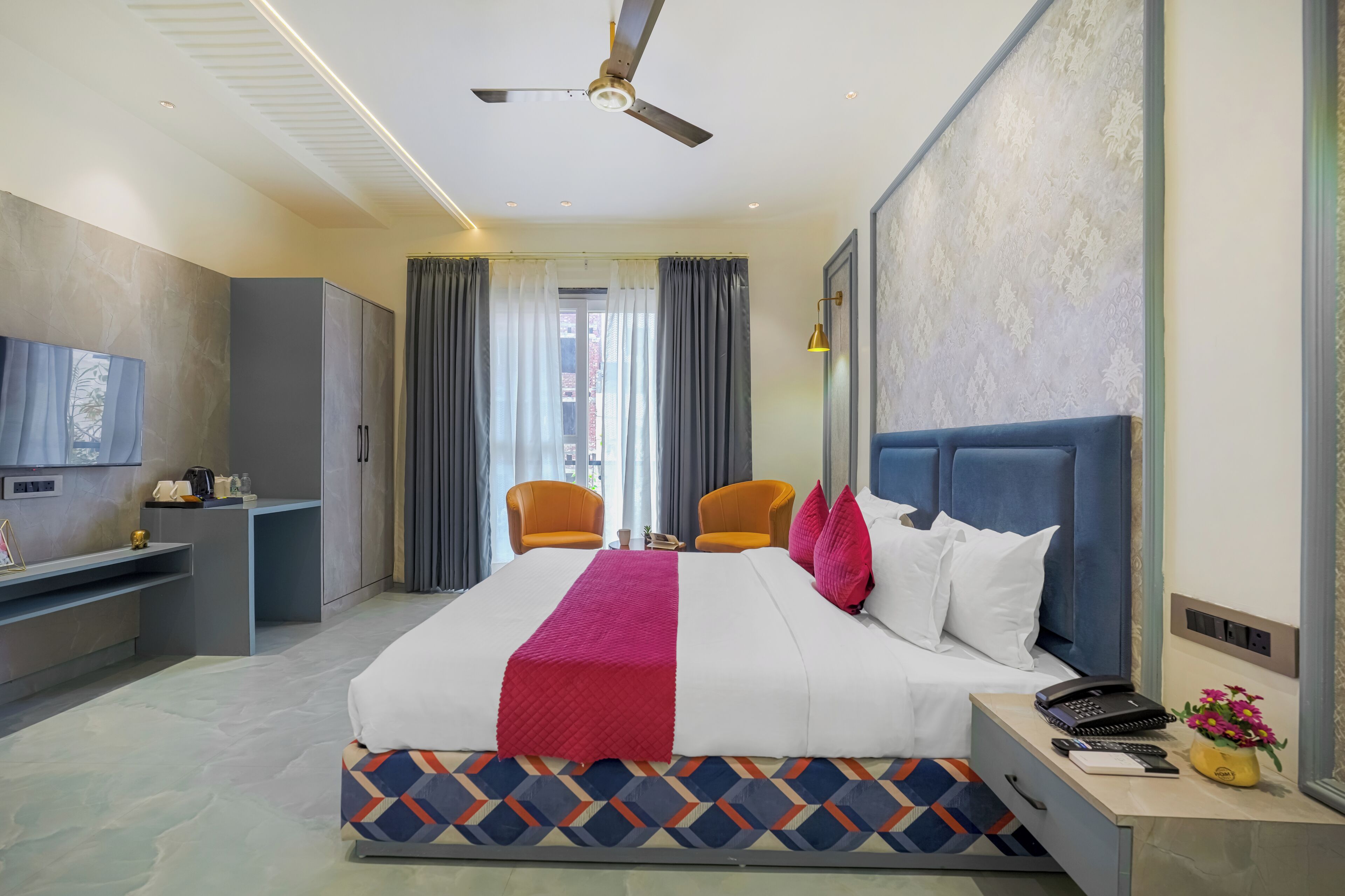 Una habitación de hotel con cama, escritorio, silla, televisión, ventilador de techo y ventana con cortinas.