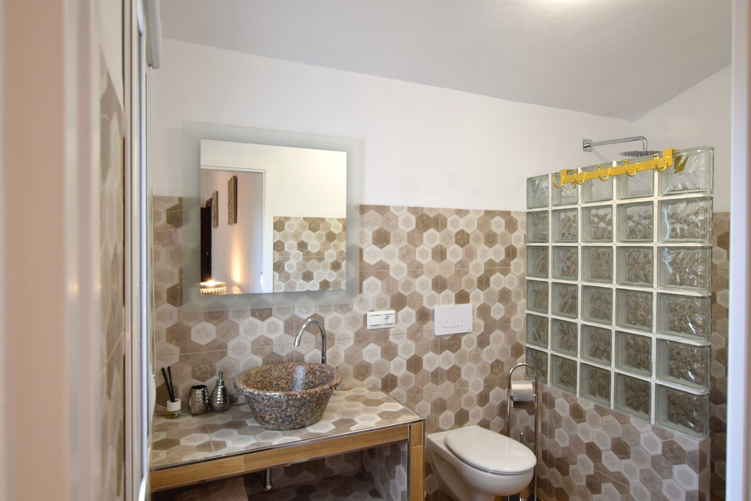 A bathroom with a hexagonal patterned tile wall, a glass shower enclosure, a wooden vanity, and a white toilet.