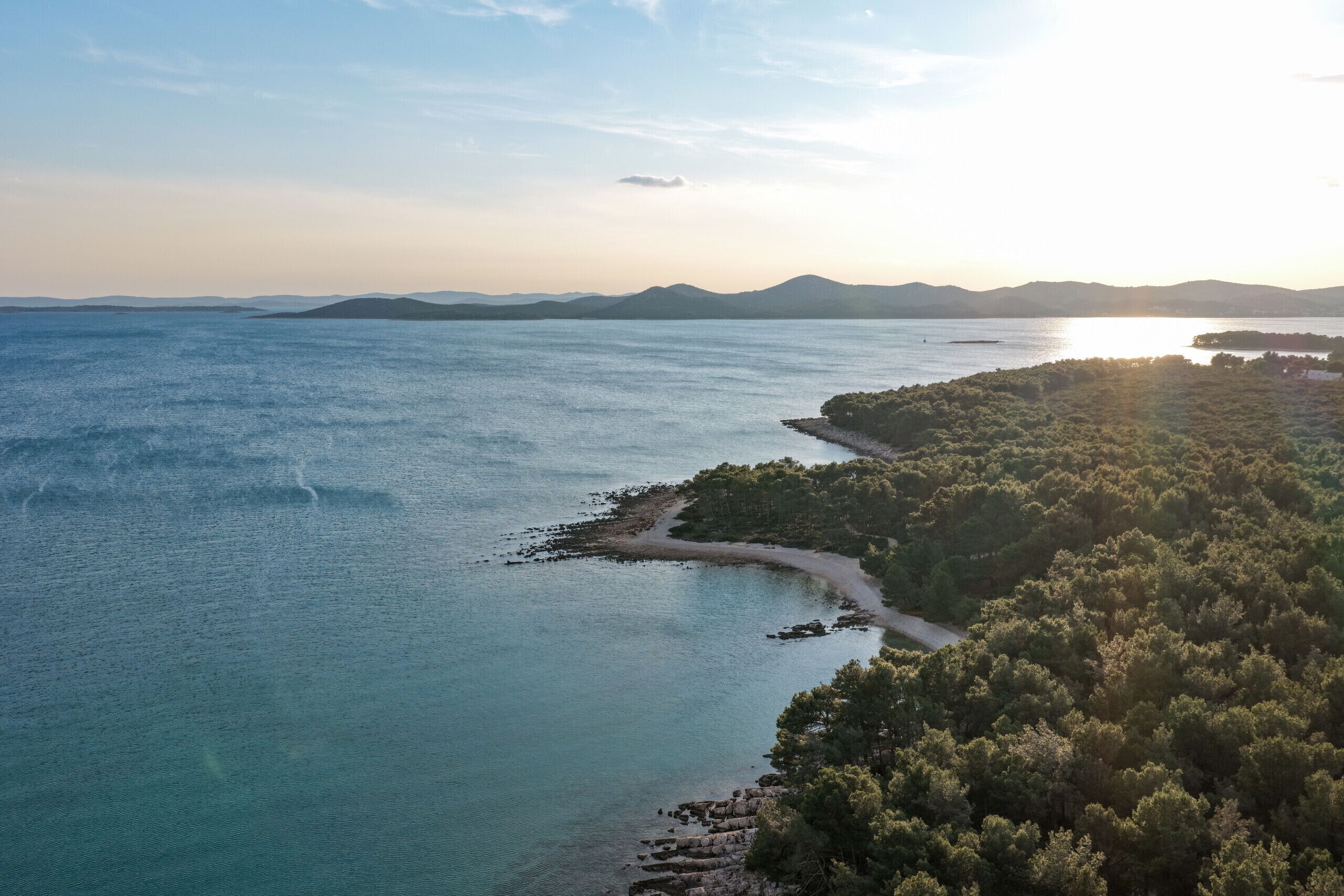 An aerial view of a coastal area with a clear path leading to a beach, surrounded by dense greenery and distant mountains.