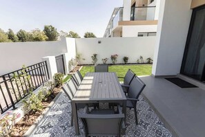 Eine moderne Außenpatio mit Holztisch und -stühlen, umgeben von einem gemusterten Boden, einem kleinen Garten und einer weißen Mauer.