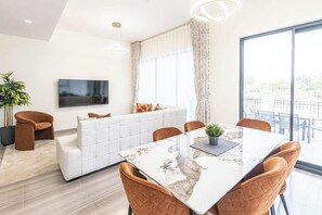 Una sala de estar moderna con un sofá blanco, una mesa de comedor con sillas, un televisor de pantalla plana montado en la pared y ventanas grandes con cortinas.