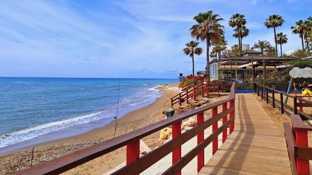 A wooden boardwalk leading to a beach with palm trees and a building with a red roof. - Beachfront studio (Calahonda)