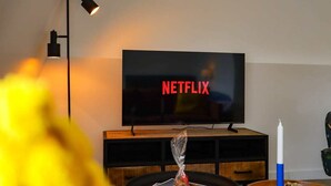 Uma sala de estar com uma televisão exibindo o logotipo da Netflix, um abajur preto, um móvel de entretenimento de madeira e uma vela sobre uma mesa.