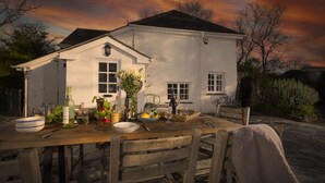 A dining table set outdoors with a white building in the background. - Sparnal Stitch nestled within the trees, just moments from sandy beaches (Rock)