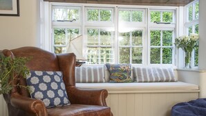 Uma poltrona de couro marrom com almofadas estampadas, um banco branco com almofadas listradas e uma janela com vista para o verde.