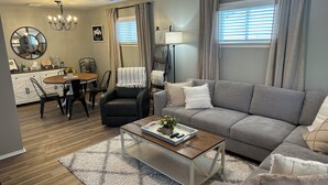 Una sala de estar moderna con un sofá gris, un sillón negro y una mesa de centro de madera. Hay un comedor con una mesa redonda y sillas.