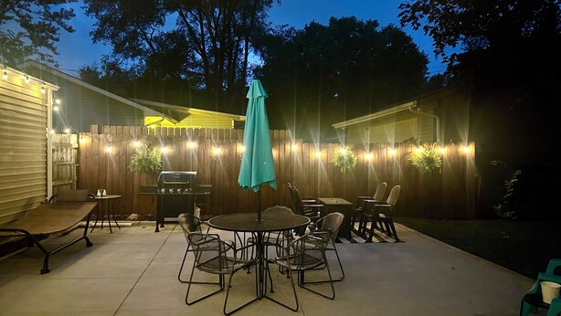 Un patio exterior con una mesa redonda y sillas, una parrilla y luces colgantes a lo largo de una valla de madera.