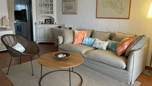 Een woonkamer met een bank, een rieten stoel, een bijzettafeltje en een kast met glazen deuren.