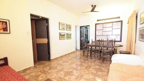 A hotel room with a bed, a desk, and a dining table with chairs. There is a door leading to another room and a ceiling fan. - Coorg Shell (Madikeri)